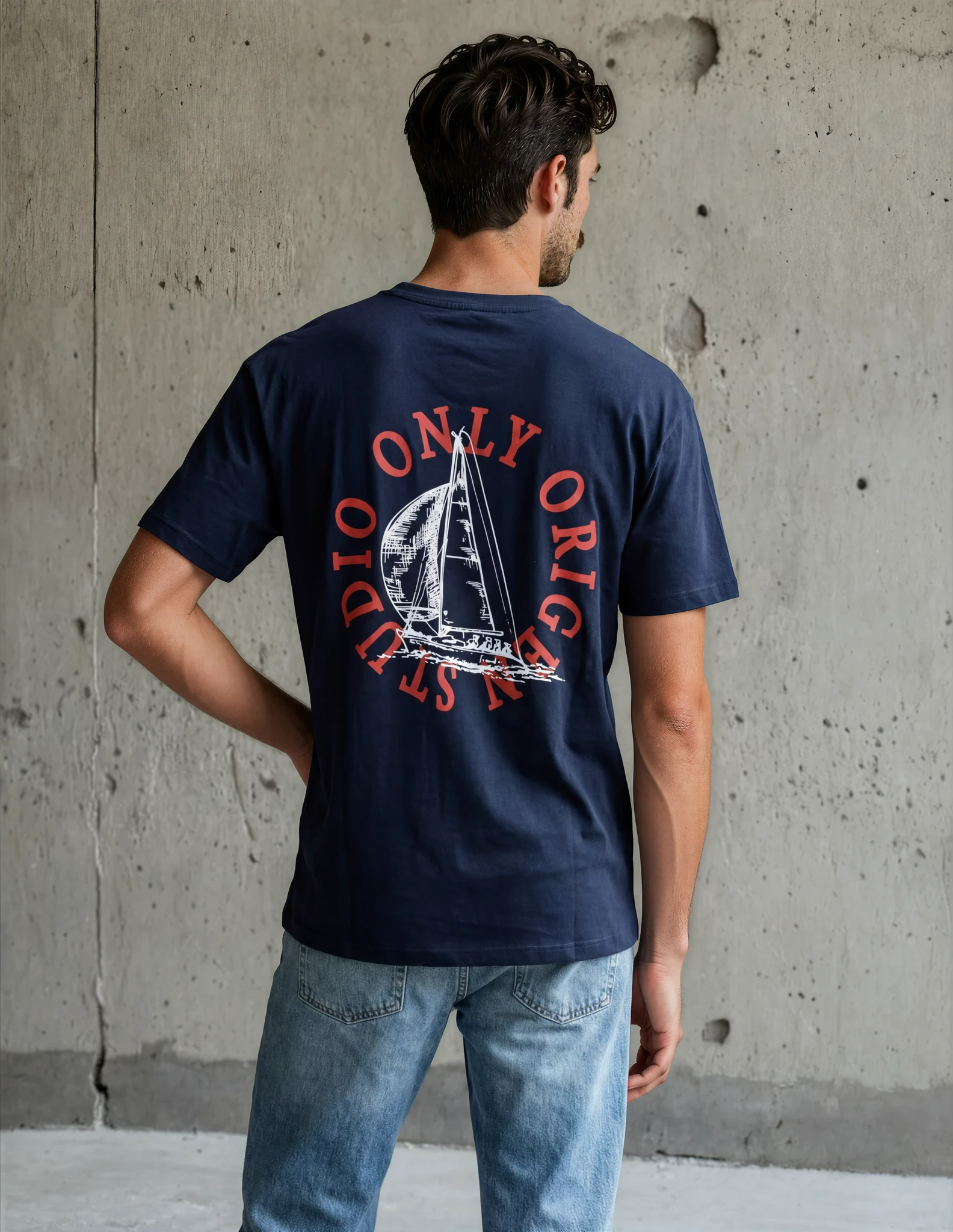 Modelo masculino de espaldas con una mano en la cadera, mostrando el ajuste y la caída de la camiseta azul marino Origen Studio y el diseño gráfico de velero en la parte posterior.