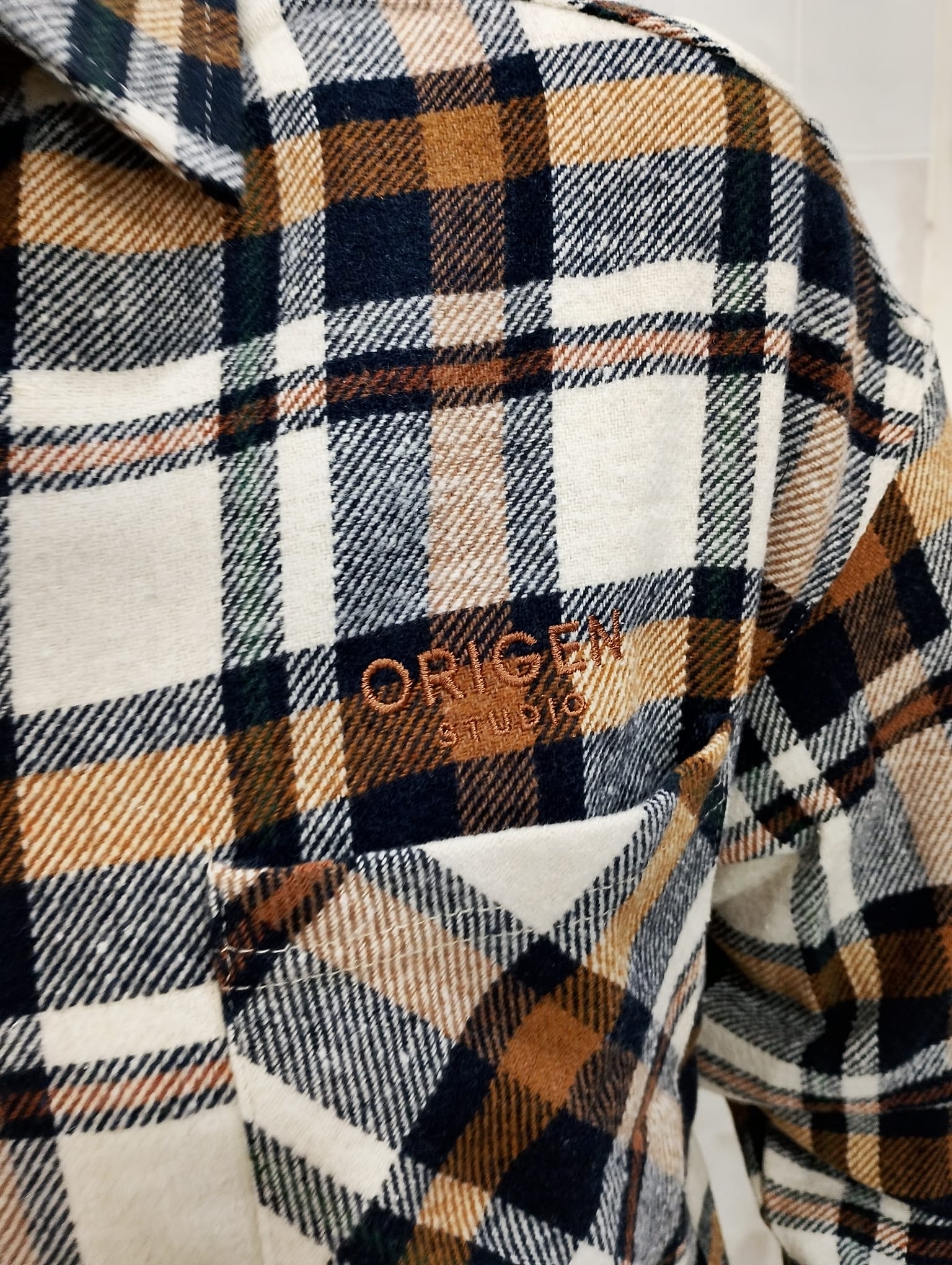 Detalle del bordado Origen Studio en el bolsillo de la sobrecamisa de cuadros beige y caramelo